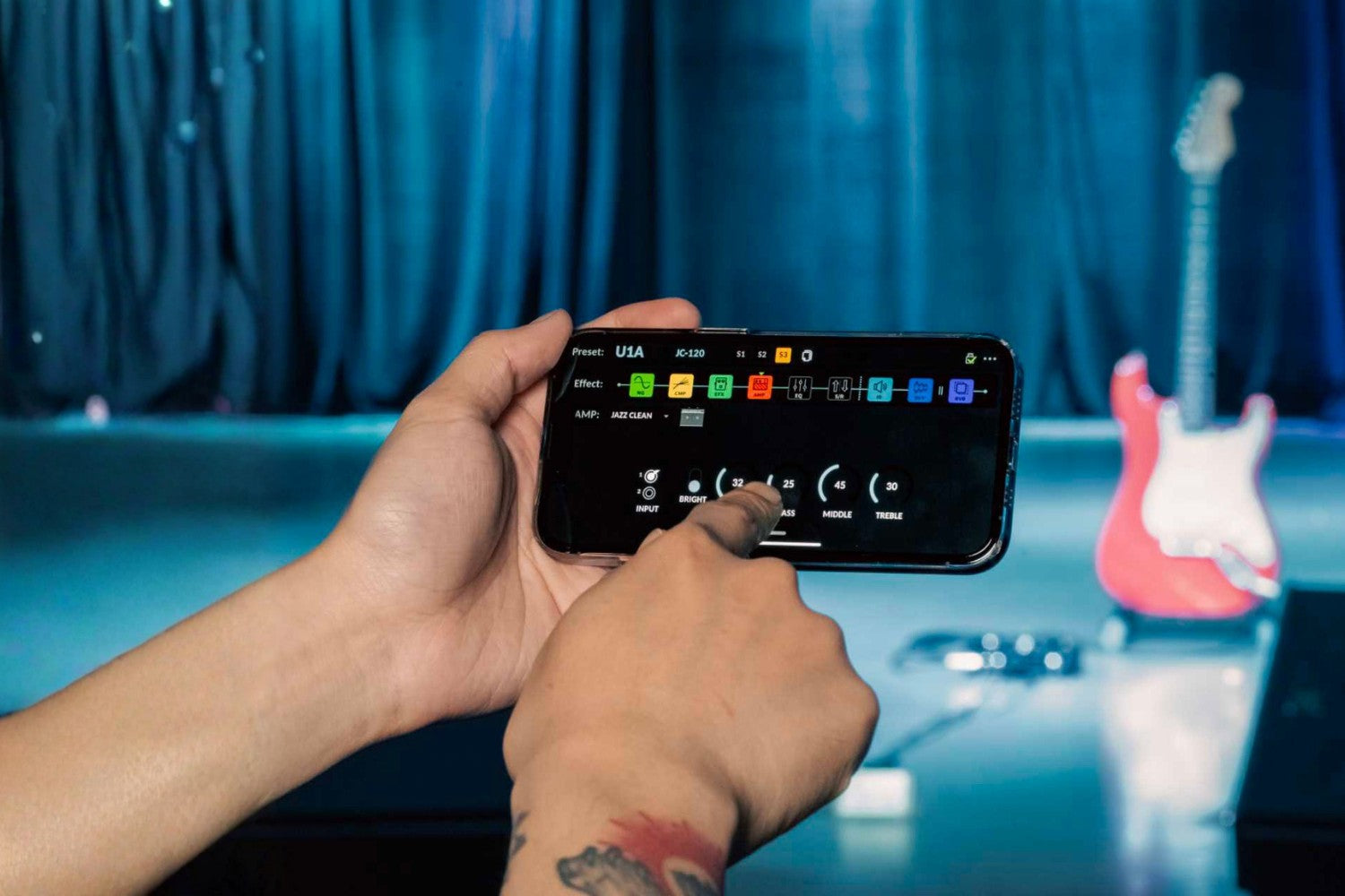 Person holding a smartphone with a music app interface in front of a guitar and curtain background