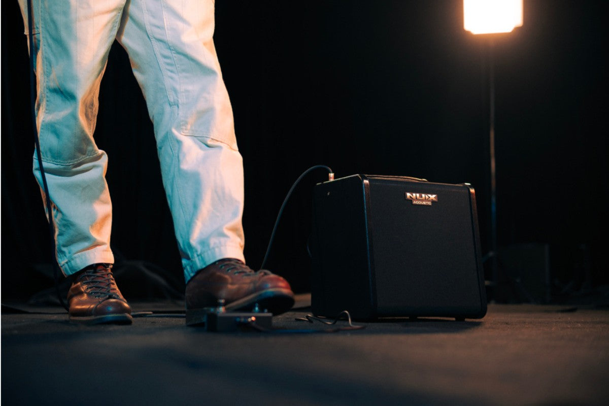 Person standing next to a small black NUX amplifier on a stage with a spotlight.