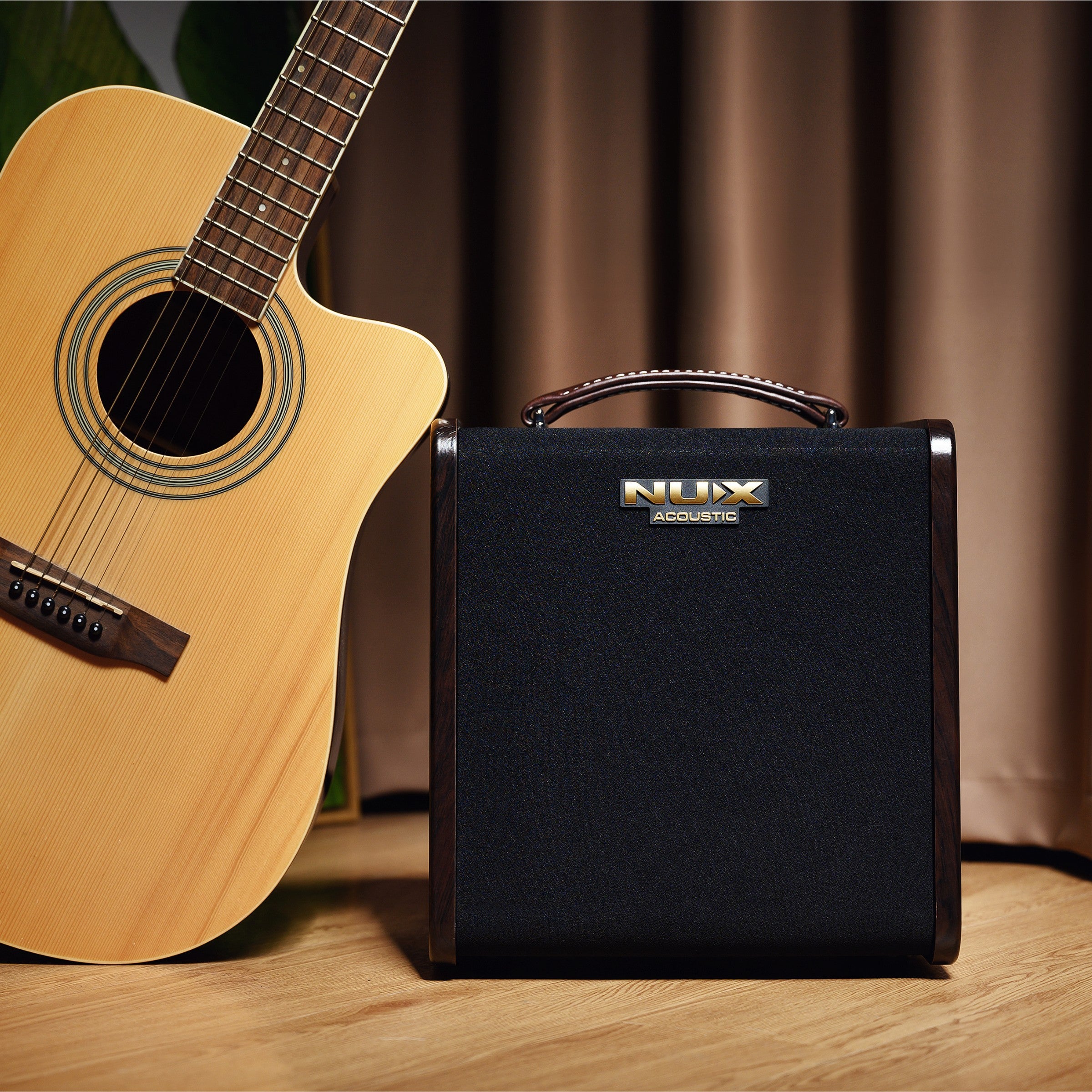 Acoustic guitar next to a NUX amplifier on a wooden surface with a blurred curtain background