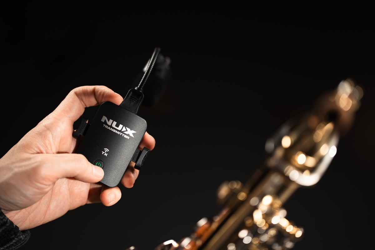 Hand holding a NUX B-6 PRO wireless transmitter with a blurred saxophone in the background