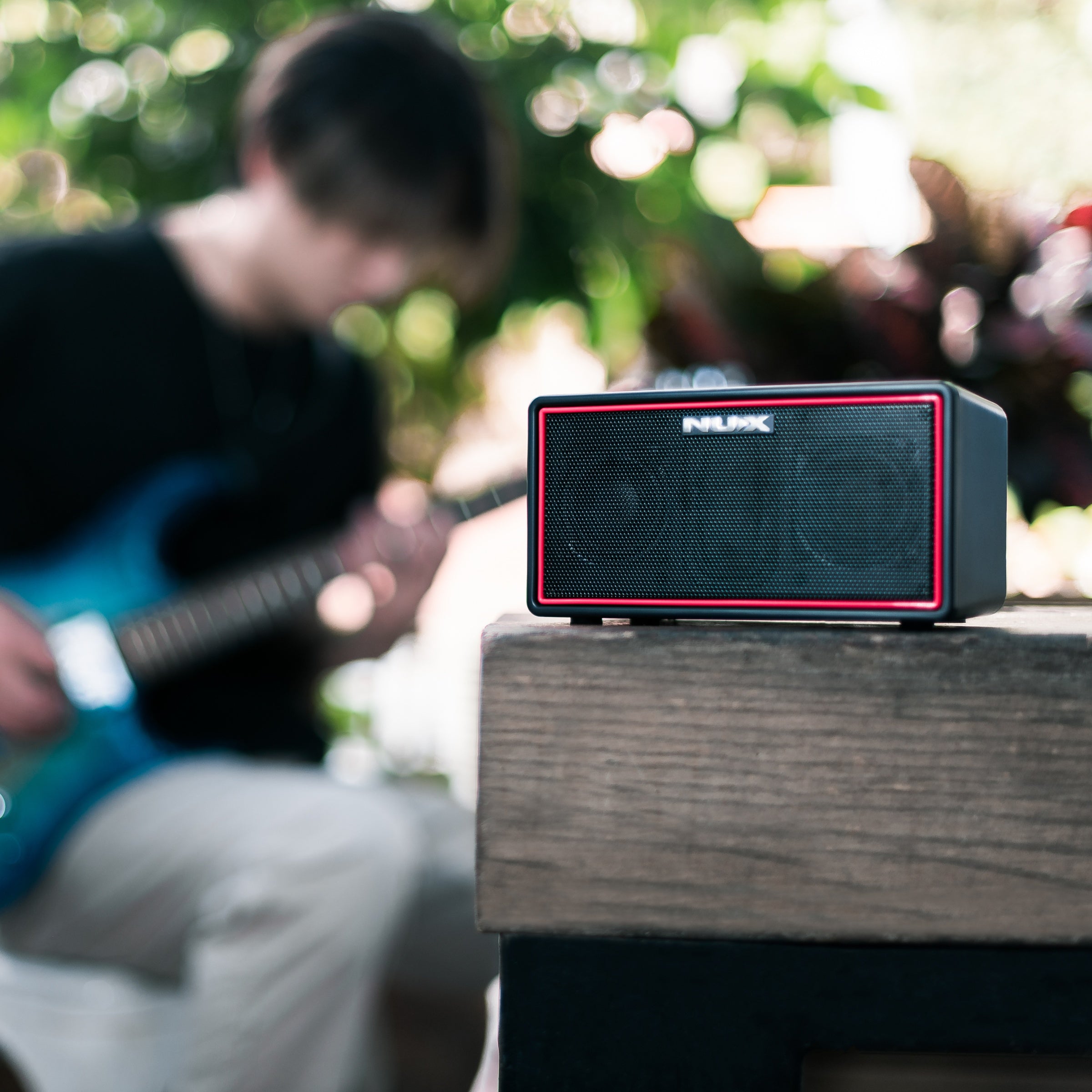 Portable speaker with red accents on a wooden surface, blurred background of people playing guitar