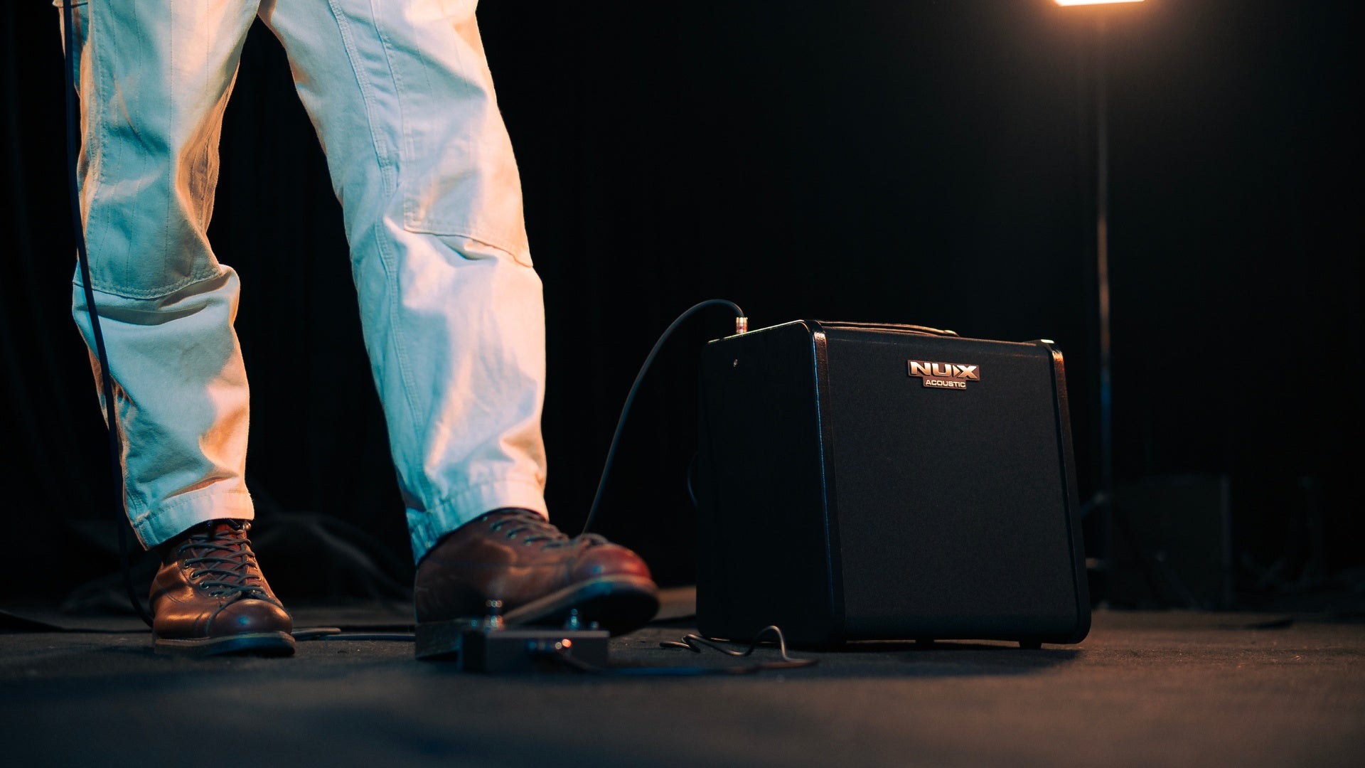 Person standing next to a black NUX amplifier with foot on a footswitch on a stage with a spotlight.