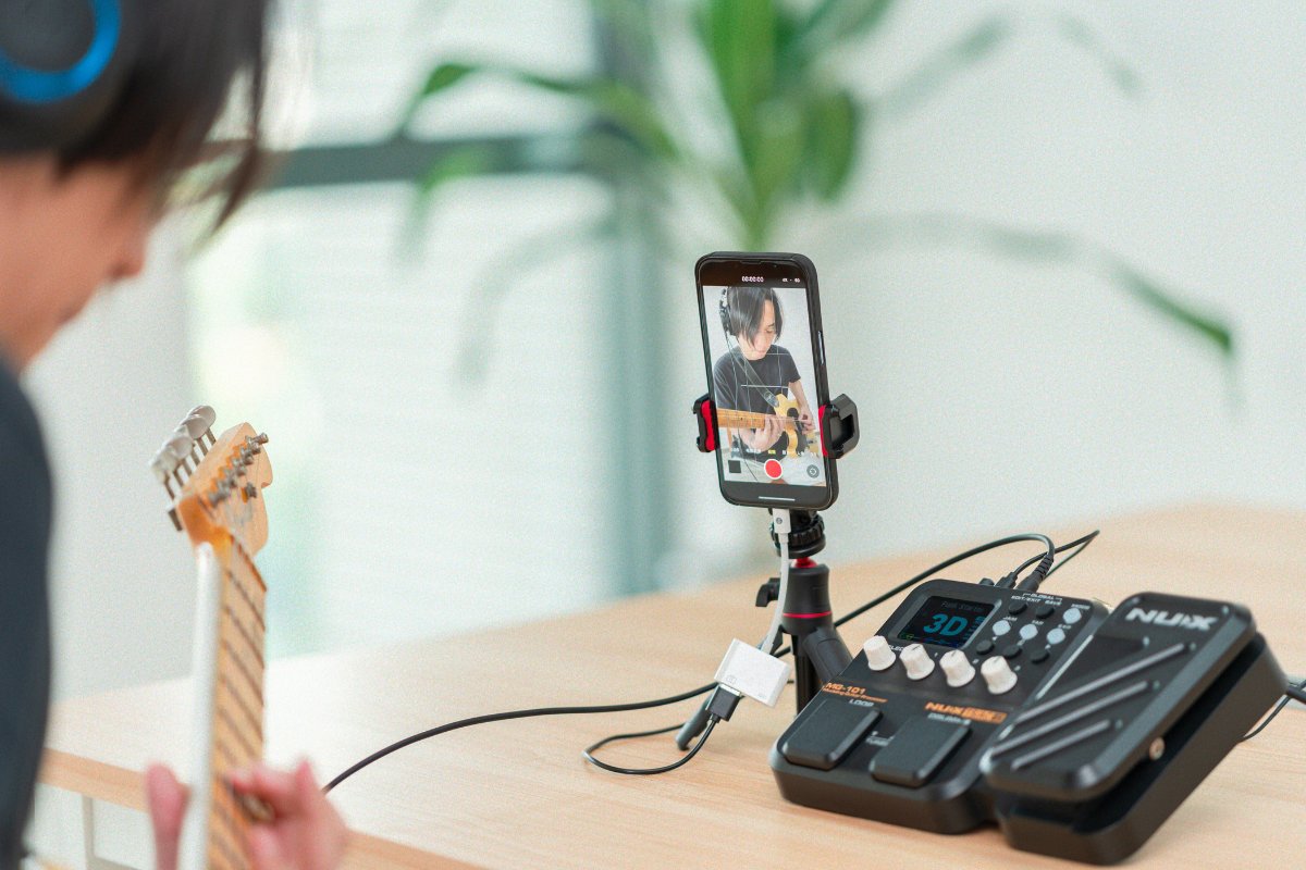 Person playing guitar livestreaming with a smartphone on a stand and a NUX mg-101 effects pedal on a table.