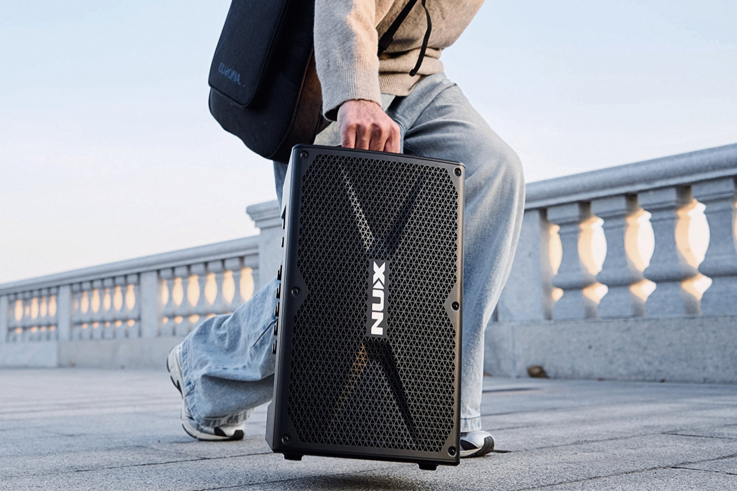 Person holding a black NUX amplifier outdoors on a bridge