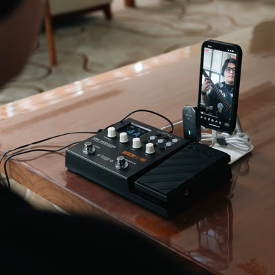 NUX MG-300 Guitar effects pedal on a wooden table with a smartphone displaying a livestream of a person playing guitar.