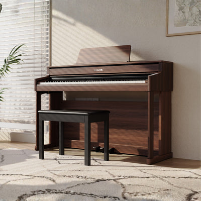 Wooden NUX upright digital piano with bench in a room with light-colored walls and a window.