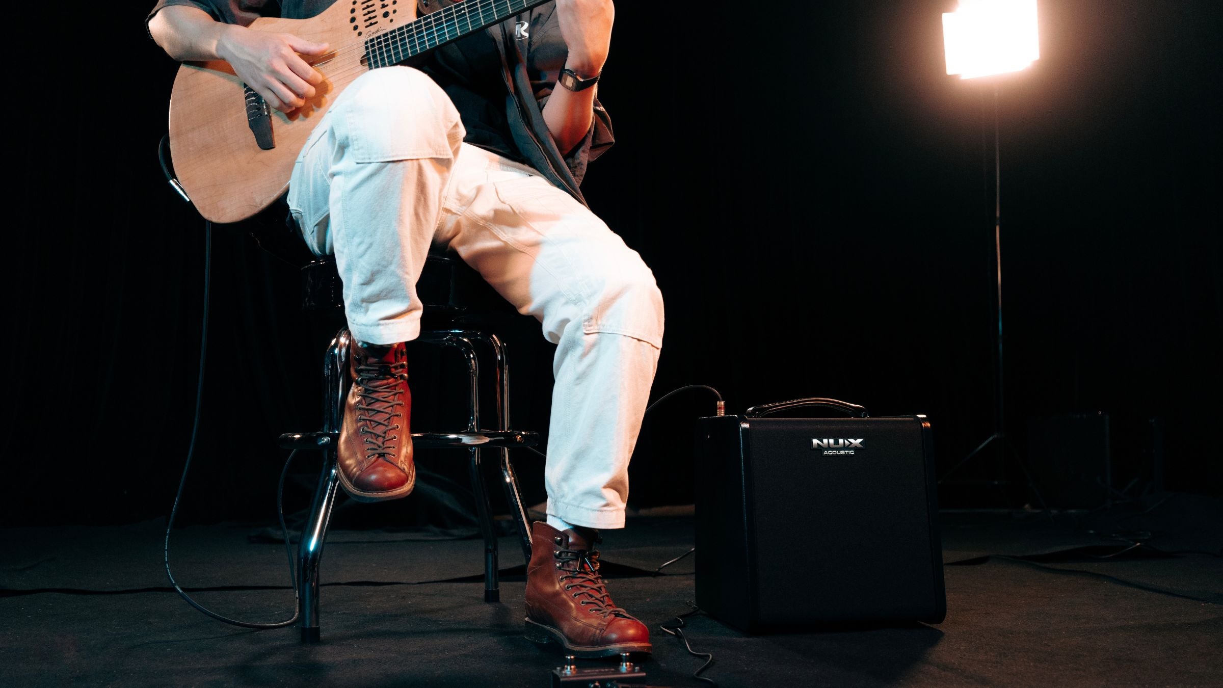 Person playing guitar on a stool with an NUX amplifier and light in the background